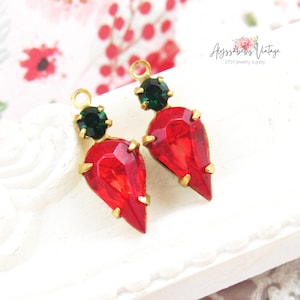 May include: Two jewelry charms featuring a faceted, pear-shaped red crystal set in a gold-toned metal prong setting, topped with a smaller round dark green crystal. Each charm includes a small circular loop at the top for attachment purposes.