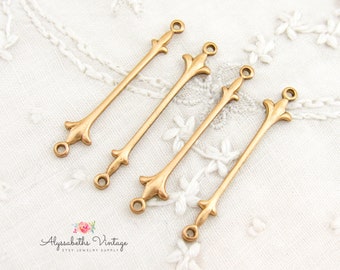 Puede incluir: Cuatro conectores de joyería de metal dorado antiguo. Cada conector tiene una barra larga y delgada con elementos decorativos en forma de flor en cada extremo y un pequeño lazo para unir a otros componentes. Los conectores se muestran sobre una superficie blanca texturizada.