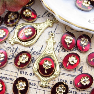 May include: A collection of jewelry components featuring two ornate gold-tone metal pendants and multiple oval-shaped cabochons. Each piece displays a deep red background with a gold-tone four-leaf clover emblem centered with a clear rhinestone.