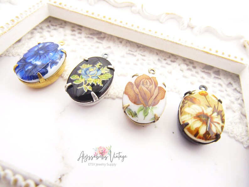 May include: Four oval-shaped porcelain pendants with floral designs. The pendants are in shades of blue, black, brown, and white. Each pendant has a gold or silver metal frame and a loop for hanging.