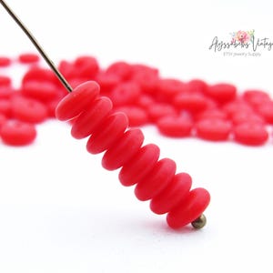 May include: Close-up of a strand of bright red, flat, disc-shaped beads. The beads are strung on a thin wire, with a brass-colored bead at the end. The background is filled with more of the same red beads, out of focus. The image is on a white background.
