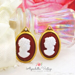 May include: Two gold-toned oval cameo pendants with a white cameo profile image on a red background.