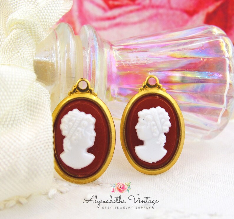 May include: Two gold-toned oval cameo pendants with a white cameo profile image on a red background.