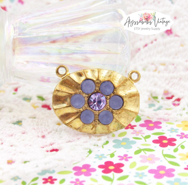 May include: Gold-toned metal pendant with a flower design featuring six purple glass stones and a smaller purple rhinestone in the center. The pendant has two jump rings for attaching to a chain or cord.