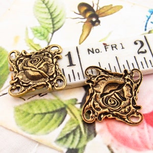 May include: Two gold-tone metal rose-shaped charms with decorative details. Each charm has three holes for attaching to jewelry or other crafts. The charms are shown next to a ruler with the measurement "1" inch.