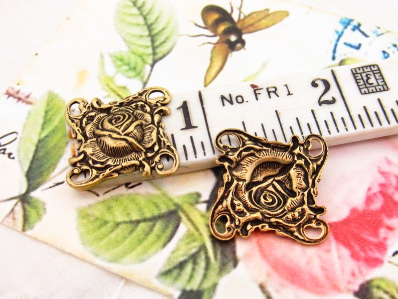 May include: Two gold-tone metal rose-shaped charms with decorative details. Each charm has three holes for attaching to jewelry or other crafts. The charms are shown next to a ruler with the measurement "1" inch.
