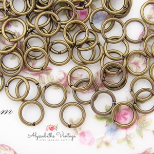May include: A collection of antique brass-colored jump rings, each approximately 6mm in diameter, scattered across a floral patterned surface. The rings are open, ready for use in jewelry making or crafts. The text "6mm" is visible.