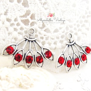 Op de afbeelding: Twee zilverkleurige, waaiervormige sieradencomponenten met helderrode kristallen accenten. Elk stuk heeft een lus aan de bovenkant voor bevestiging. De achtergrond is een witte kanten stof. De tekst "Alyssabeths Vintage" is zichtbaar.