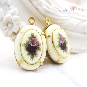 May include: Two gold-tone oval-shaped pendant charms with white enamel and a floral design. The floral design features pink and purple roses with green leaves.