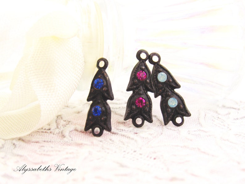 Puede incluir: Dos abalorios de metal de estilo antiguo en forma de flor con tres piedras de cristal cada uno. Los abalorios tienen forma de flor con tres p&eacute;talos. Las piedras de cristal son azules, rosas y azul claro.