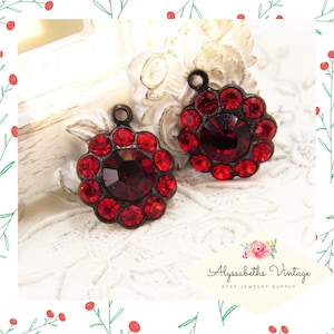 May include: Two red rhinestone embellishments with a black metal setting and a loop for hanging. The rhinestones are arranged in a circular pattern around a larger, central rhinestone.