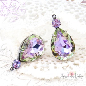 Pode incluir: Dois brincos de cristal em forma de lágrima com tons iridescentes verdes e roxos. Cada brinco apresenta um cristal redondo menor de cor roxo claro.