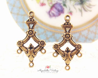 Puede incluir: Dos hallazgos de pendientes de metal dorado con un diseño decorativo que incluye una corona, flores y bucles. Los hallazgos están sobre una superficie blanca.