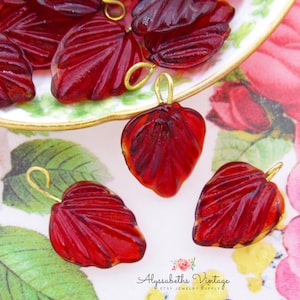 May include: A group of red glass leaf shaped charms with gold jump rings. The charms are on a white plate with a gold rim and a floral pattern.