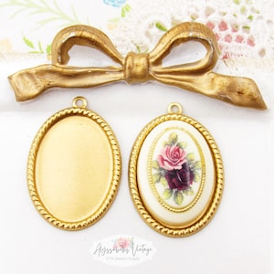 May include: Gold-toned vintage jewelry findings, including two oval pendant blanks and one with a floral rose design. A gold bow-shaped embellishment is also present. The items are displayed on a white surface.