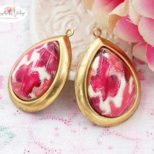 May include: Two gold-tone teardrop-shaped pendant charms with a pink and white marbled design. The charms have a loop at the top for attaching to jewelry.