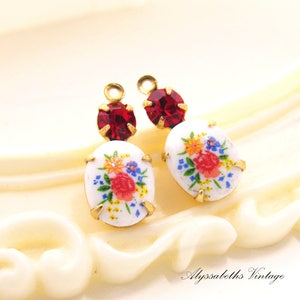 May include: A pair of gold-tone earrings with white oval-shaped glass charms featuring a floral design and red rhinestone accents.