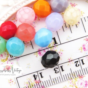 May include: Assortment of faceted glass beads in various colors, including pink, red, orange, blue, green, purple, and black. The beads are arranged on a white surface with a ruler for scale.