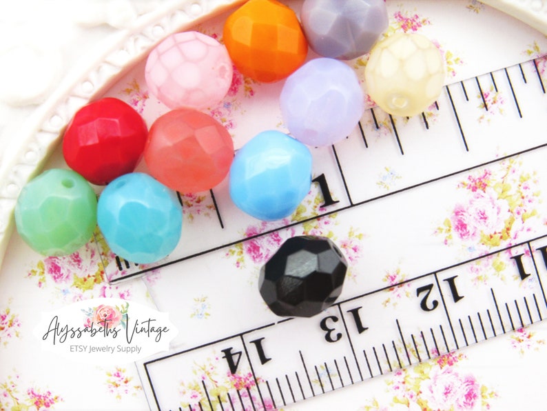 May include: Assortment of faceted glass beads in various colors, including pink, red, orange, blue, green, purple, and black. The beads are arranged on a white surface with a ruler for scale.