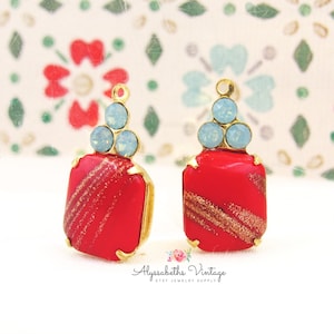 May include: Two red rectangular jewelry components with gold accents and light blue crystal embellishments. Each component has a gold-colored loop at the top and a diagonal gold glitter design. The components are from Alyssabeths Vintage.