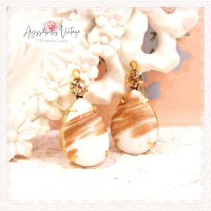 May include: A pair of white teardrop-shaped earrings with gold accents and a brown swirl pattern. Each earring has a small clear rhinestone at the top.