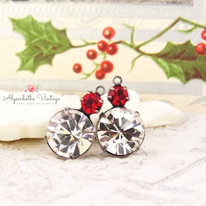 May include: Two vintage-style earrings featuring large, clear, round crystals with a smaller, red, round crystal accent. The earrings have a silver-tone metal setting. The background includes a holly and berry design, suggesting a holiday theme.