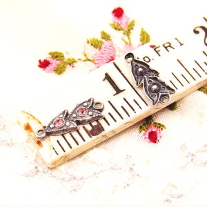 May include: Two silver-toned metal charms with pink rhinestone accents. The charms are shaped like a stylized leaf with a loop at the top for attaching to a necklace or bracelet. The charms are shown on a white fabric background with a ruler.