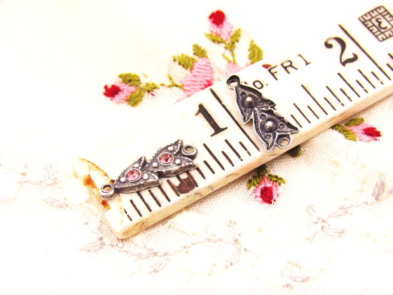 May include: Two silver-toned metal charms with pink rhinestone accents. The charms are shaped like a stylized leaf with a loop at the top for attaching to a necklace or bracelet. The charms are shown on a white fabric background with a ruler.