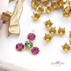 May include: A collection of gold-toned metal jewelry findings in a three-pronged star shape. One piece is set with three round faceted rhinestones, featuring two vibrant magenta stones and one central light green stone, while others remain empty.