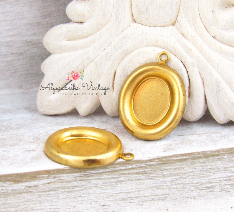 May include: Two gold-toned metal oval pendant trays with a raised edge and a loop for hanging. The trays are blank and ready for embellishment.