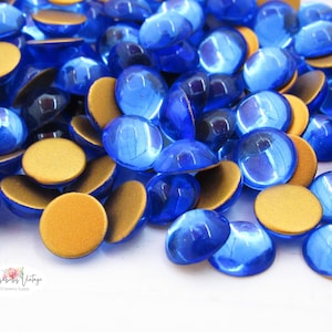 Könnte beinhalten: Eine Ansammlung runder Bastelverzierungen im Cabochon-Stil mit einer durchscheinenden, leuchtend blauen gewölbten Oberseite und einer flachen, mattgoldenen Unterseite. Die Teile liegen verstreut in einem Haufen und zeigen ihre zweifarbige Optik.