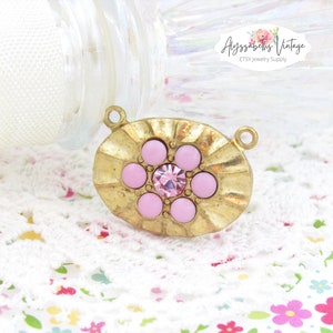 May include: Gold-toned oval pendant with a raised, textured design. The pendant features a central pink rhinestone surrounded by six smaller pink rhinestones.