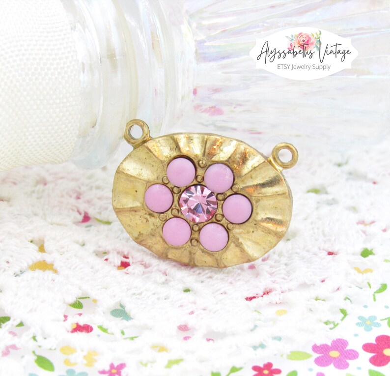 May include: Gold-toned oval pendant with a raised, textured design. The pendant features a central pink rhinestone surrounded by six smaller pink rhinestones.