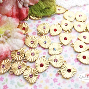 May include: Gold heart-shaped charms with a textured surface and a single rhinestone in the center. The rhinestones are in shades of pink and red.