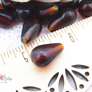 Peut inclure: Gros plan sur des perles en verre brun en forme de larme, mesurant environ 3,8 cm de long, sur un fond blanc avec une r&egrave;gle pour l'&eacute;chelle.