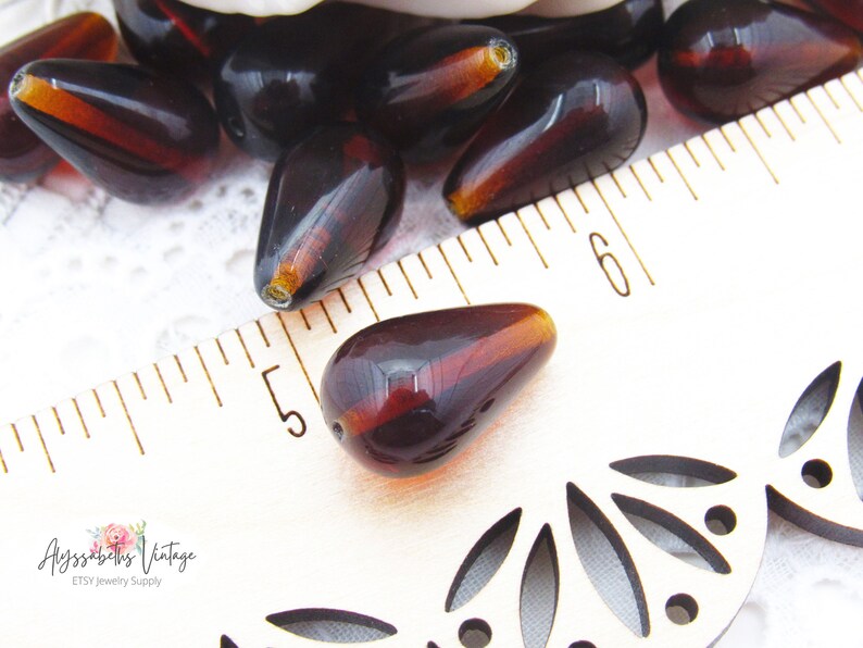 Peut inclure: Gros plan sur des perles en verre brun en forme de larme, mesurant environ 3,8 cm de long, sur un fond blanc avec une r&egrave;gle pour l'&eacute;chelle.