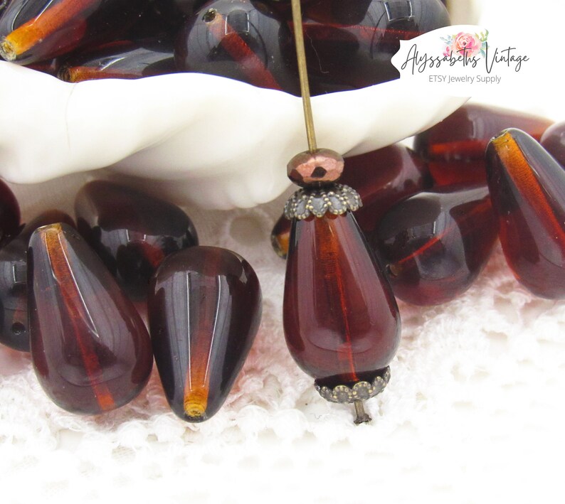 Peut inclure: Un bol de perles en verre rouge fonc&eacute; en forme de larme avec une perle en m&eacute;tal dor&eacute; sur une &eacute;pingle. Les perles sont sur une surface en dentelle blanche. Le texte "Alyssabeths Vintage ETSY Jewelry Supply" est en arri&egrave;re-plan.