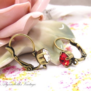 May include: A pair of antique gold hoop earrings with a clear crystal and a red crystal. The earrings are on a white background with a pink rose.