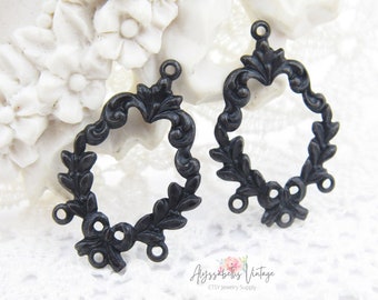 May include: Two black metal ornate oval-shaped frames with three jump rings each. The frames have a leaf and scroll design.