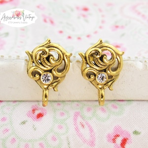 May include: Two ornate, gold-colored jewelry findings with a swirling design and a clear, round rhinestone in the center. Each finding has a small loop at the bottom. The background is a floral pattern.