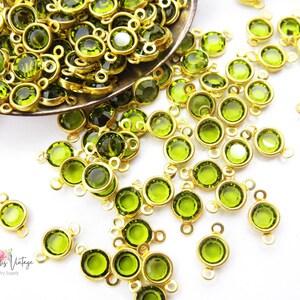 May include: A pile of green glass beads with gold-toned metal loops. The beads are scattered on a white surface.