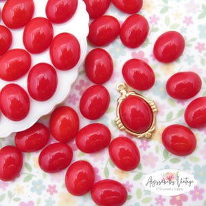 May include: A collection of red oval-shaped cabochons, some are on a white plate and others are scattered on a floral patterned surface.  The cabochons are smooth and glossy.