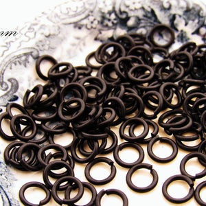 May include: A close-up of a pile of black metal jump rings, 6mm in diameter. The rings are on a white surface with a decorative pattern.