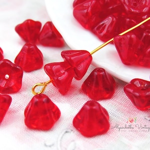 May include: A scattering of red glass flower beads, some with five points and others with six points, are displayed on a white surface. The beads are translucent and have a slight sparkle. A gold wire is threaded through some of the beads.