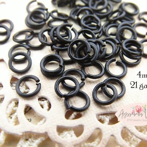 May include: A close-up of a pile of 4mm black jump rings, 21 gauge. The rings are on a white lace background. 4mm 21 gauge