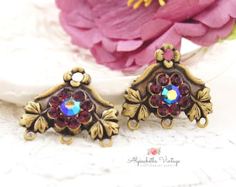 May include: Two antique brass-toned jewellery components. Each features a central iridescent crystal surrounded by smaller red crystals, set within a floral design. The components have leaf accents and small loops for attachment, ideal for vintage-inspired jewellery.