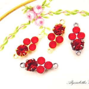 May include: Four red rhinestone jewelry components with gold and silver-tone metal findings. Each component features a large, round red rhinestone surrounded by smaller red rhinestones. The components are displayed on a white surface with floral accents.