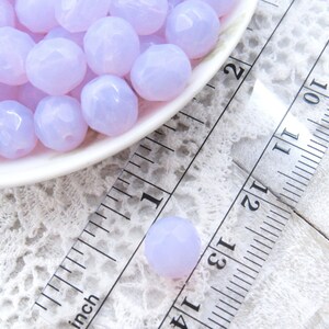 May include: A close-up of a bowl filled with lavender-colored faceted glass beads. The beads are approximately 1/2 inch in diameter and are arranged in a random pattern. The bowl is sitting on a white lace tablecloth next to a ruler.