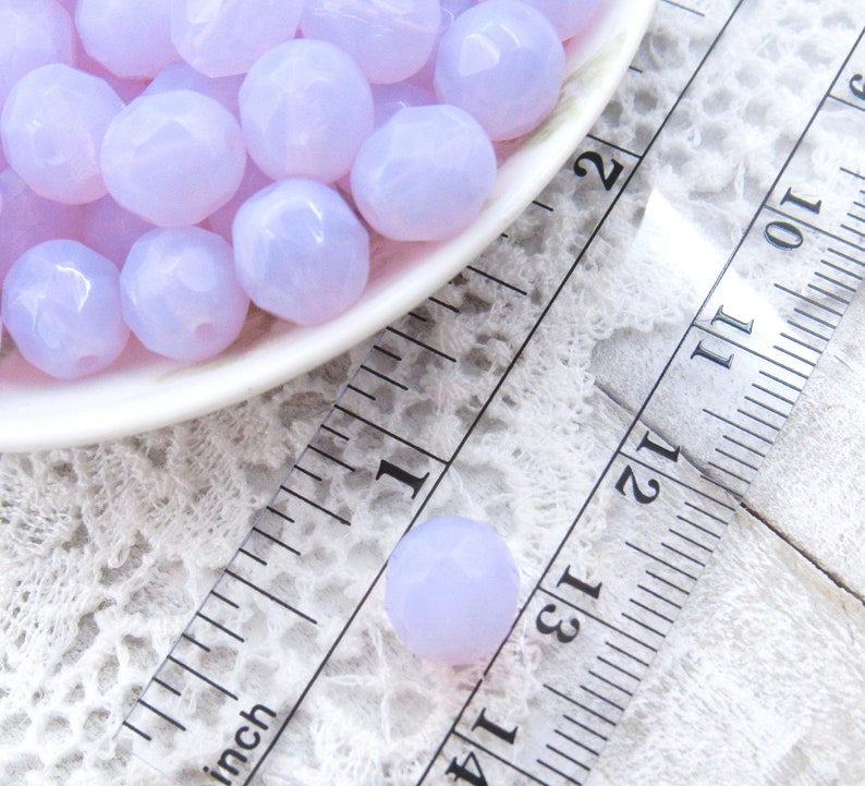 May include: A close-up of a bowl filled with lavender-colored faceted glass beads. The beads are approximately 1/2 inch in diameter and are arranged in a random pattern. The bowl is sitting on a white lace tablecloth next to a ruler.