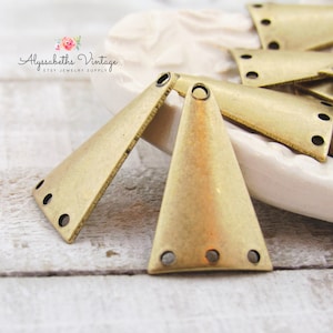 May include: Two gold-toned metal triangle-shaped pendant charms with four holes each. The charms are on a white surface.